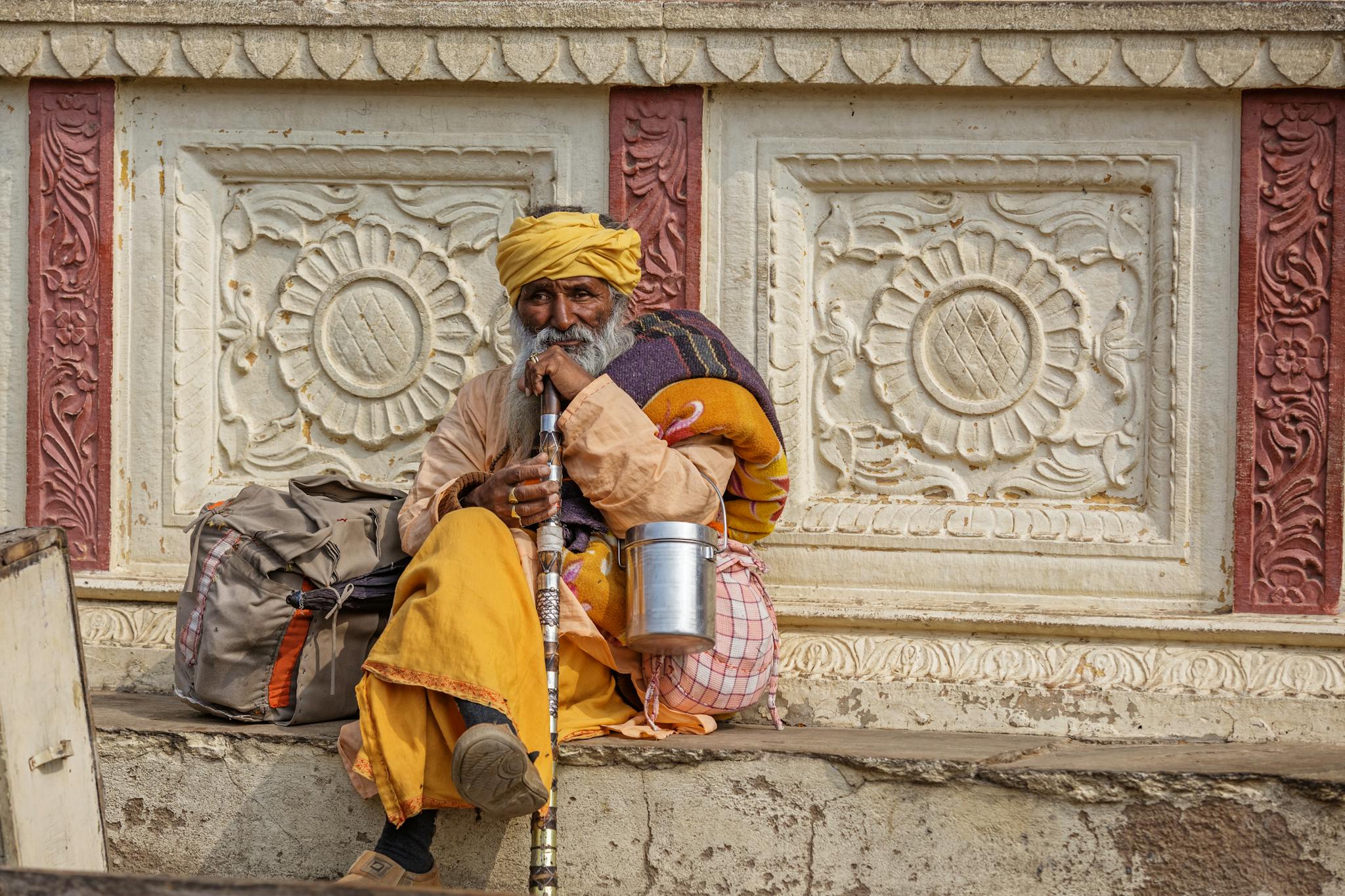 Full body of elderly Indian man in colorful turban sitting with belongings and ornamental cane on street