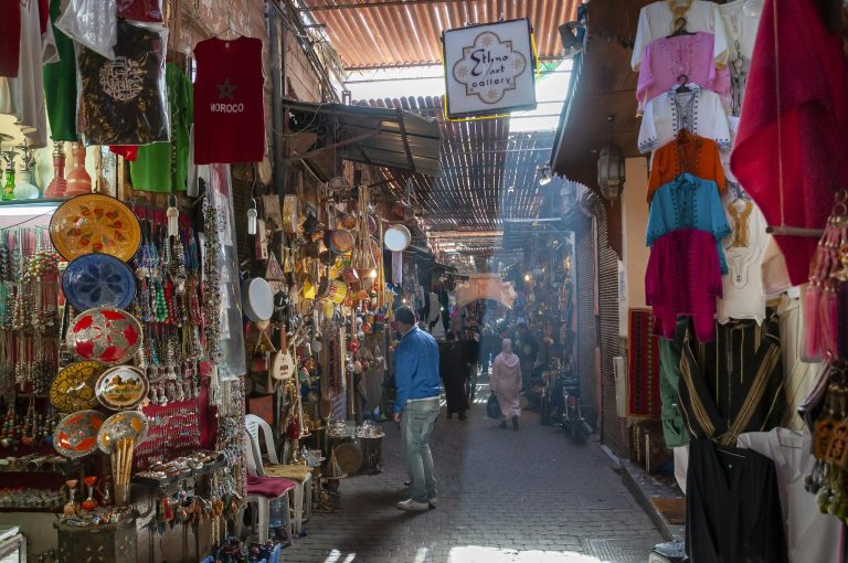 Unrecognizable people walking in local bazaar near stalls with various goods and souvenirs and traditional clothes on sunny day in Marrakesh